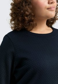 Schwarzer Sweatshirt mit Struktur, runder Ausschnitt und abgerundeten Schulternähten, mit einem subtilen, ineinandergreifenden Muster über dem Stoff.