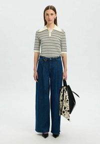 Kvinna i en kräm- och svartrandig kortärmad skjorta, vida blå jeans, svarta spetsiga skor, bär en svart väska med en prickig scarf.