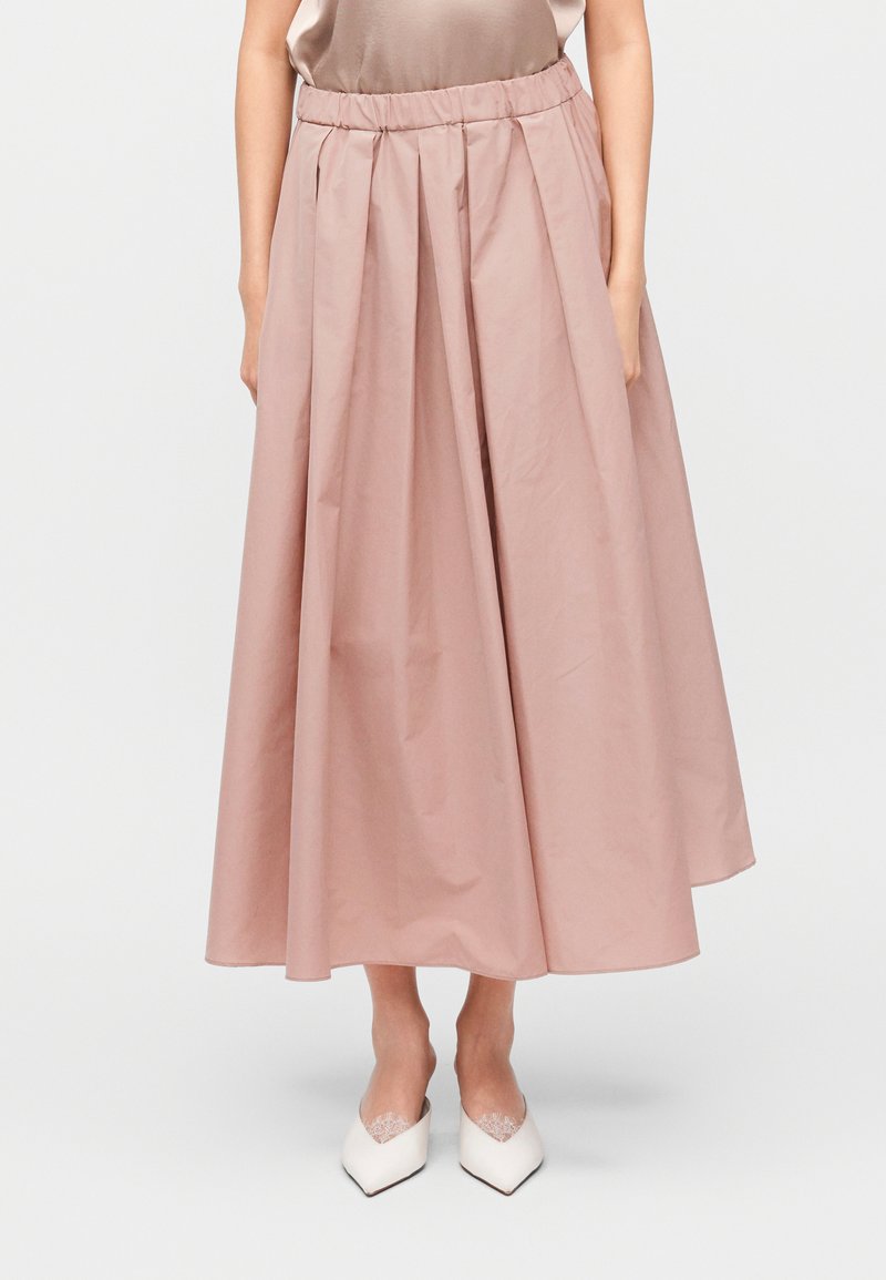 Ροζ midi φούστα με πιέτες και ελαστική μέση, κατασκευασμένη από λείο ύφασμα, σε συνδυασμό με λευκά παπούτσια με μυτερή μύτη. Απλός και μινιμαλιστικός σχεδιασμός.