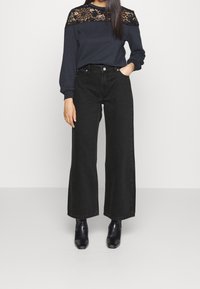 Mustat leveälahkeiset farkut, joita on yhdistetty tummansiniseen yläosaan, jossa on pitsiyksityiskohta hartioissa. Sileä kangas, pitkät hihat ja istuvat hihansuut.