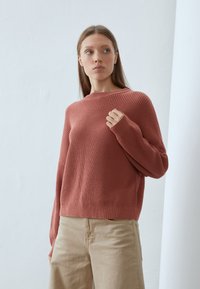Rust-colored sweater with a ribbed texture, crew neckline, and drop shoulder sleeves. Paired with light khaki pants, displayed against a white background.