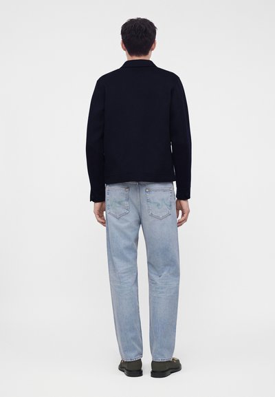 Homme debout de dos, portant une veste noire, un jean bleu clair et des chaussures foncées sur un fond blanc uni.