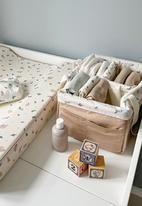 Koszyk na tkaniny wypełniony różnorodnymi miękkimi materiałami, kwiecistą podkładką do przewijania, beżową butelką z balsamem oraz drewnianymi blokami w podstawowych kolorach.