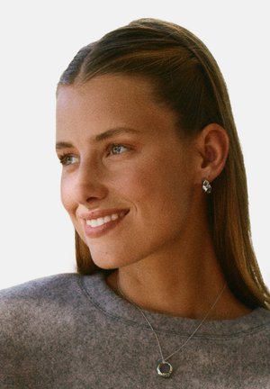 Orecchini a bottone in argento e una collana con pendente circolare su un maglione grigio chiaro. I capelli sono lisci e il soggetto ha un'espressione neutra.