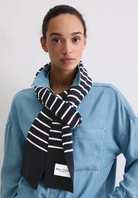 Écharpe à rayures noires et blanches en tissu doux, avec une extrémité unie noire portant une étiquette de marque, portée sur une chemise en jean.