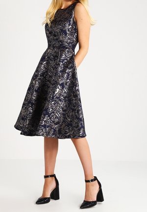 Femme portant une robe sans manches bleu marine avec un motif floral argenté, des poches et des talons blocs noirs à bride à la cheville, debout sur un fond blanc uni.