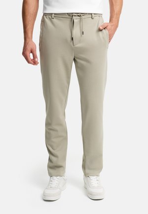 Man draagt beige broek met trekkoord en witte sneakers, staand met één hand in de zak tegen een effen achtergrond.