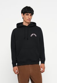 Sort hættetrøje med en frontlomme, snørelukning i hætten og broderet "Tommy Jeans" logo i hvid og pink. Blød stofstruktur.
