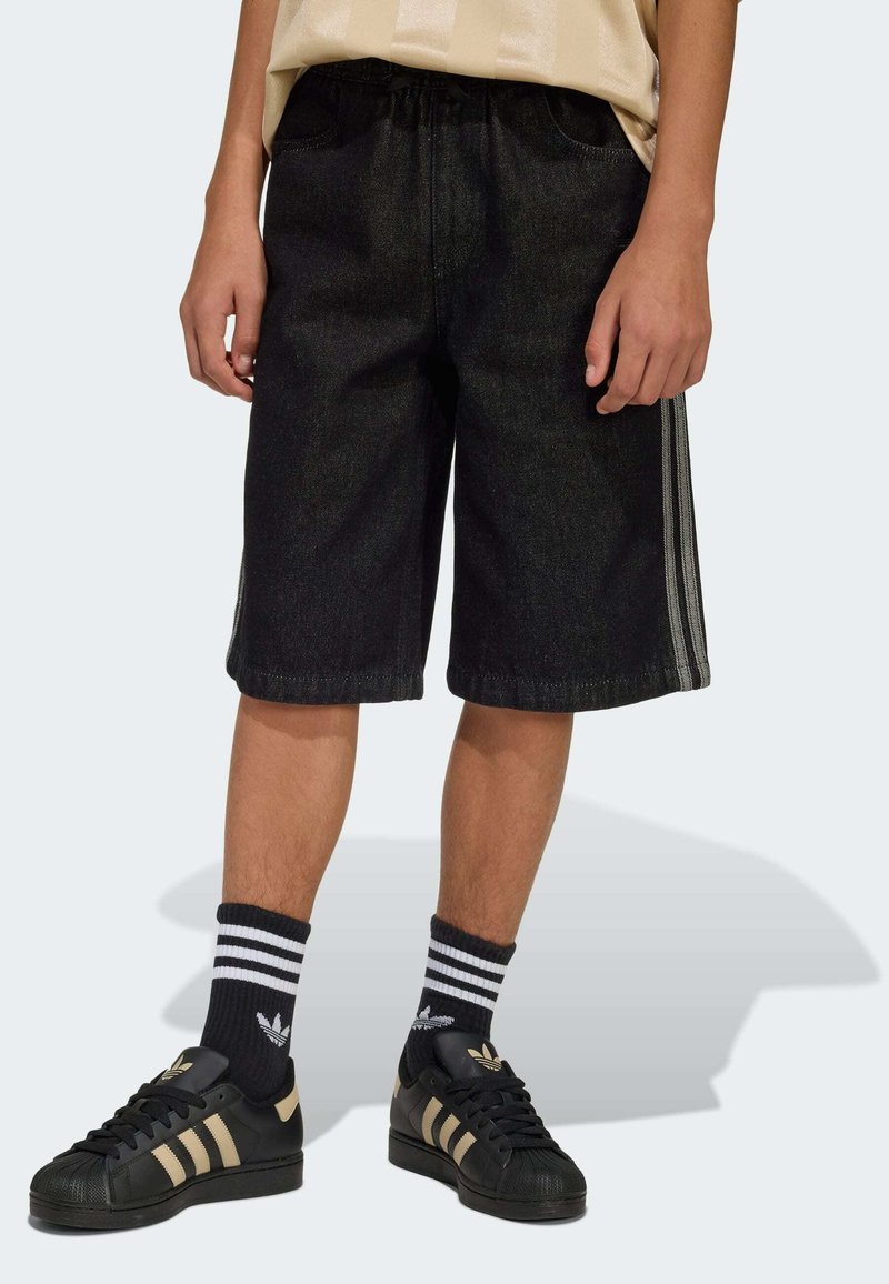 Shorts in denim nero con vestibilità morbida, caratterizzati da strisce laterali. Abbinati a sneakers nere e calzini a righe, evidenziando uno stile casual.