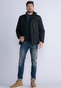 Giacca nera isolata con tasche a bottoni, colletto rialzato e dettagli con zip, indossata sopra un maglione blu navy, abbinata a jeans blu e stivali beige.