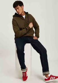 Oliwkowa bluza z kapturem na zamek z czarną podszewką, noszona na białej koszulce; niebieskie jeansy; czerwone sneakersy Adidas z białymi paskami.