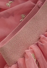 Pink mesh fabric features a soft, textured surface with a gold-threaded band and ruffled accents, creating layered detail and visual interest.