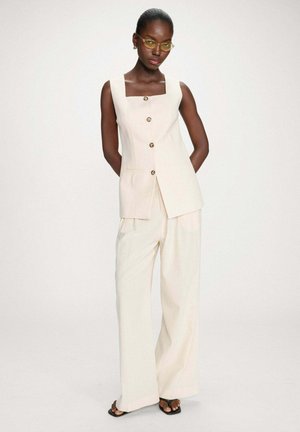 Femme portant un gilet sans manches crème boutonné, un pantalon large assorti, des lunettes à verres jaunes et des sandales noires, debout devant un fond uni.