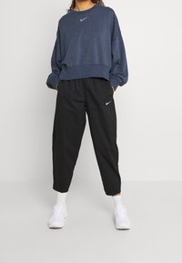 Blå sweatshirt med långa ärmar och ribbat nederkant, i kombination med svarta avsmalnande byxor. Båda har Nike-logotypen i vitt. Vita sneakers bärs.