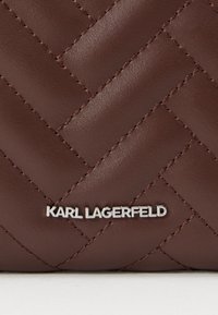 Brązowa pikowana skórzana torebka z błyszczącym srebrnym logo "KARL LAGERFELD" na dole. Teksturowana powierzchnia z przeszytymi detalami.