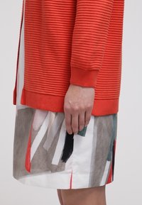 Texturerad orange kardigan med ribbad design, ihop med en mönstrad kjol med geometriska former i rött, grönt och svart på en vit bakgrund.