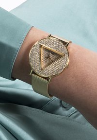 Reloj de oro con una esfera triangular incrustada de cristales; presenta una correa de malla y un sutil contraste textural con un fondo verde sedoso.