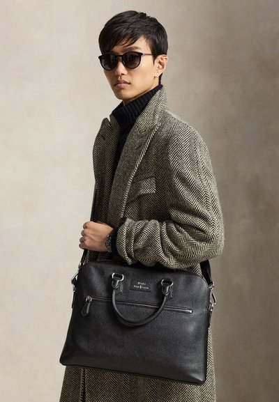 Homme portant des lunettes de soleil et un manteau à chevrons tient une mallette noire en cuir Polo Ralph Lauren avec double poignée et une poche zippée.