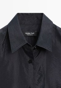 Chemise en tissu noir brillant avec col pointu et boutons visibles, affichant une étiquette Massimo Dutti à l'intérieur de l'encolure.