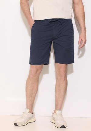 Homme portant un short bleu marine, des baskets beiges et un t-shirt beige clair, debout avec une main dans la poche contre un mur blanc.