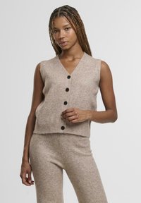 Béžový pletený bezrukávový svetr s výstřihem do V, s pěti černými knoflíky a žebrovaným lemem, v kombinaci s odpovídajícími texturovanými kalhotami.