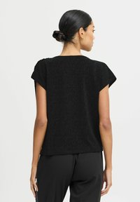 Top nero testurizzato con maniche corte, caratterizzato da una vestibilità ampia e un collo rotondo. Indossato con pantaloni neri, mostrato da dietro.