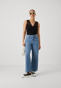 Haut noir avec dentelle, jeans larges taille haute bleu clair, baskets noires avec semelles blanches et sac à main clair, portés par une silhouette debout.