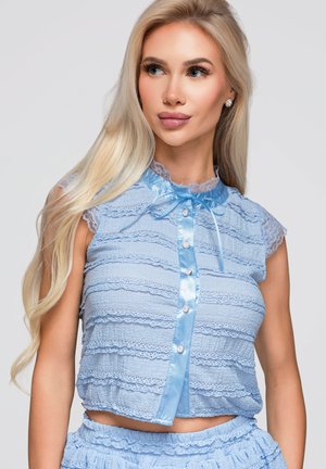 Femme blonde portant un haut en dentelle sans manches bleu clair avec des boutons en satin et un nœud, regardant légèrement sur le côté sur un fond neutre.
