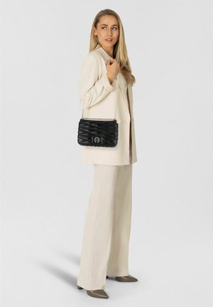 Sac à main noir matelassé avec une bandoulière chaîne, présentant un accent logo subtil. Le modèle porte un blazer crème et un pantalon large.