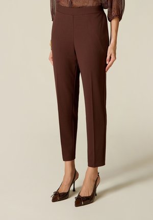 Kvinde iført mørkebrune, slim-fit ankel-lange bukser og mørkebrune højhælede sko med spidse tæer og sløjfer.