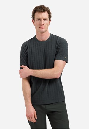 Man met krullend haar die een getextureerd donkergrijs shirt met korte mouwen en een donkere broek draagt, staand met één arm over de andere, tegen een effen lichte achtergrond.