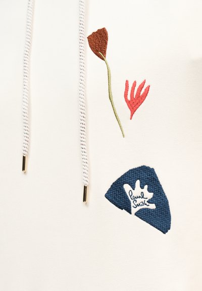 Biela tkanina s bielymi šnúrkami a farebnými vyšívanými abstraktnými tvarmi, vrátane červenej ruky, hnedej listovej a modrého tvaru s bielym detailom.