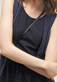Navy Kleid mit einem strukturierten Blumenmuster, geschichtet mit einer dunklen Weste. Eine dünne schwarze Umhängetasche betont das Outfit.