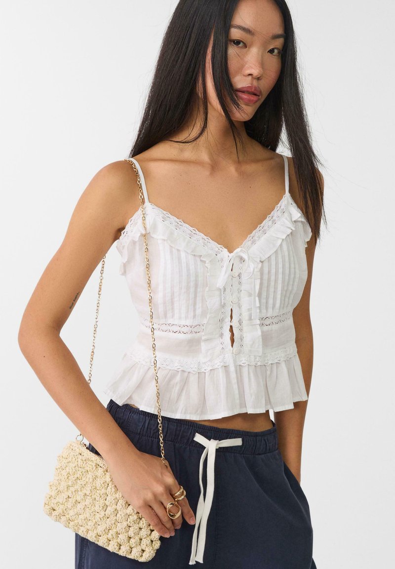 Top bianco a balze con rifiniture in pizzo e allacciatura frontale. Abbinato a una borsa croquet beige testurizzata e pantaloni blu navy. Cintura a catena argentata.