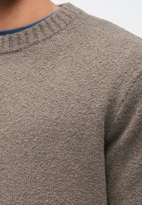 Harmaa neulepaita, jossa on teksturoitu viimeistely ja resorireunus kaula-aukossa, ja kauluksen sisäpuolella näkyy sininen kerros.