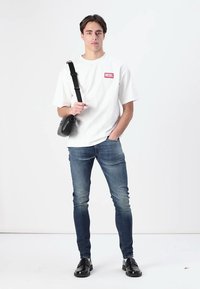 T-shirt blanc à manches courtes avec logo "DIESEL" rouge, jeans en denim bleu et sac à bandoulière noir. Le modèle porte des chaussures noires, se tenant sur un fond blanc.