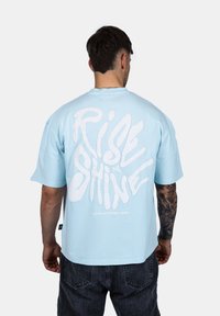 Svetle modré oversized bavlněné tričko s velkým bílým nápisem "Rise & Shine" na zadní straně, krátké rukávy a kulatý výstřih.