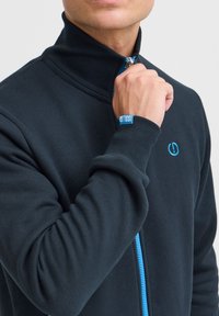 Mørkeblå hættetrøje med lynlås, høj krave, ribstrikkede ærmeafslutninger og en kontrastfarvet blå lynlås. Logodetalje på brystet og håndleddet.