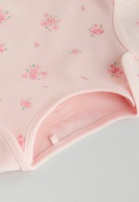 Hele roosa beebibody lilledemustriga, mis sisaldab väikseid roose ja rohelisi lehti. Pehme kangas, ümara kaelusega ja tugevalt õmmeldud.