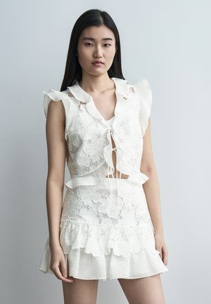 Νεαρή γυναίκα που φοράει λευκό δαντελένιο τοπ χωρίς μανίκια με βολάν δεμένο στο μπροστινό μέρος και ταιριαστή δαντελένια φούστα, μπροστά σε απλό γκρι φόντο.