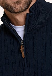 Marineblauwe gebreide trui met hoge kraag en halve rits, voorzien van een kabelbreipatroon en een bruin leren accent op de ritstrekkers.