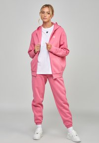 Roze rits hoodie en bijpassende joggers van zachte stof. Witte t-shirt met ronde hals eronder. Witte sneakers. Eenvoudig design, losse pasvorm.