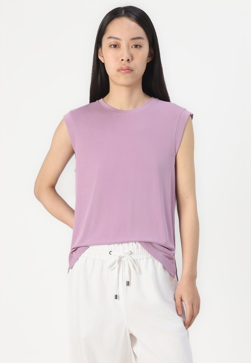 Lilafarbene ärmellose Bluse aus weichem Stoff mit Rundhalsausschnitt und lockerer Passform, kombiniert mit weißen Jogginghosen mit Kordelzug. Sanfte Textur, lässiges Design.
