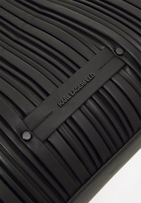 Zwarte leren tas met verticale plooien, een gladde textuur en een gemerkte schouderband met gegraveerde tekst. Decoratieve hardware is minimaal.