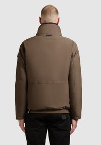 Brown padded jacket with high collar, textured fabric, and a matte finish. Features a logo tag on the collar and is paired with black jeans.