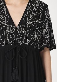 Rochie neagră cu decolteu în V, decorată cu broderie florală albă pe mâneci. Talie legată cu ciucuri, material texturat și tiv evazat.