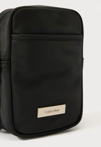 Fekete nylon válltáska lekerekített sarkokkal, felső cipzáras záródással és egy fém logó táblával az elején. Simább textúra, minimalista dizájn.