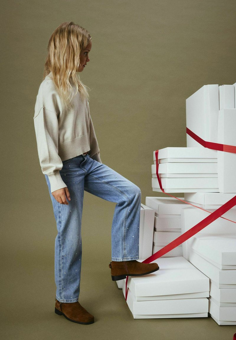 Maglione corto beige chiaro, jeans blu chiaro con piccoli punti bianchi, stivali alla caviglia marroni, in piedi accanto a scatole regalo bianche legate con un nastro rosso.