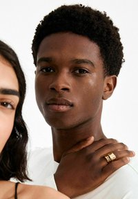 Kuldne sõrmus tekstuursete aktsentidega, kantud tumeda nahaga mehe paremal käel, valgel taustal. Kujutatud isiku särk on heledat värvi.