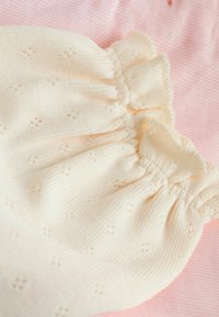 Tela beige clara con pequeños patrones de ojal bordados, con bordes fruncidos y acentos de volantes, sobre un fondo rosa suave.